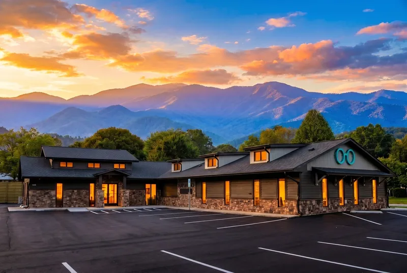 The Haywood Family Eye Care building on Sulphur Springs Road at sunset — a single-story stone-and-gable office with warm-lit windows and the teal round-glasses logo on the gable, the Blue Ridge Mountains rising behind it