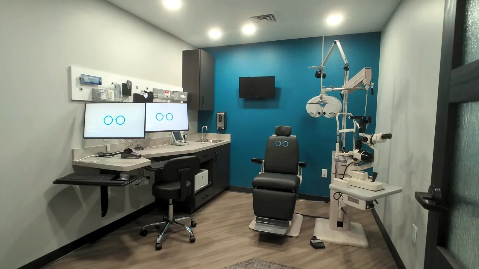 One of our exam lanes: twin monitors showing the teal glasses logo, a slit-lamp microscope and phoropter on the teal accent wall, and the patient exam chair